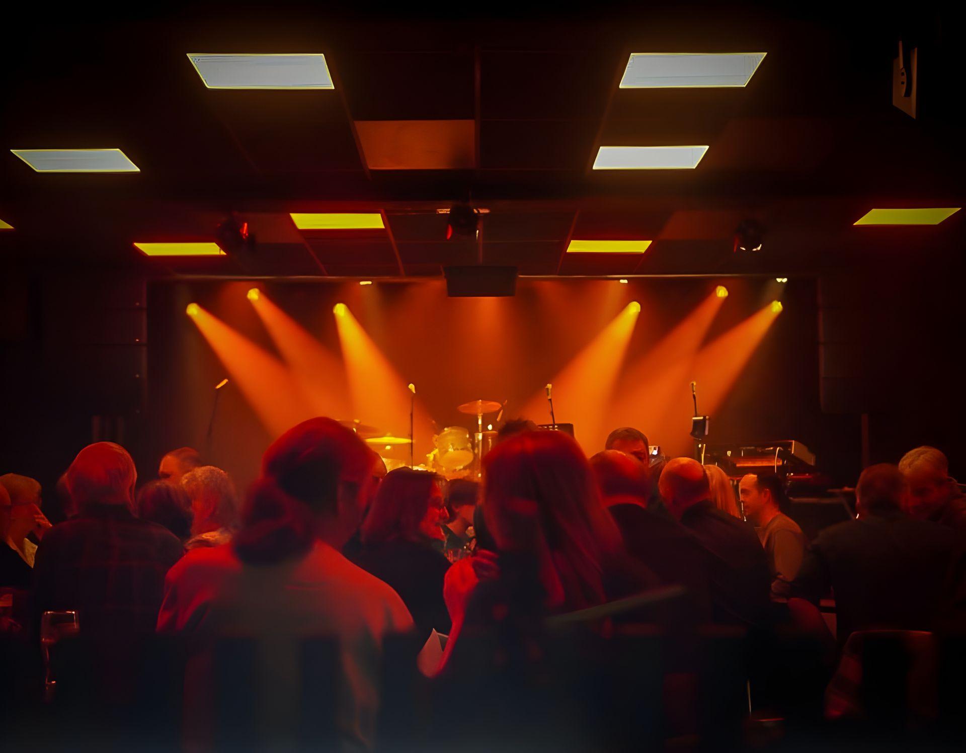 Concert venue with stage lights shining on musical instruments, viewed from a crowd of seated audience members.