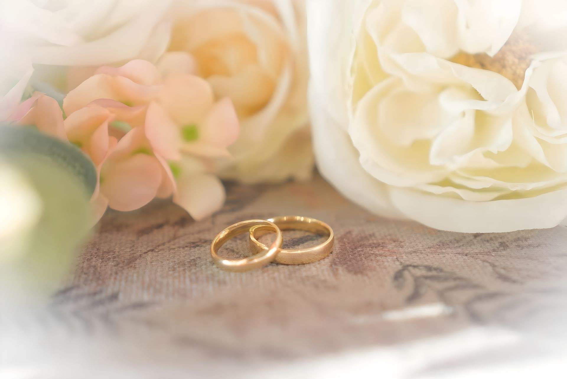 Two gold wedding rings on a textured surface surrounded by soft pink and white flowers in the background.
