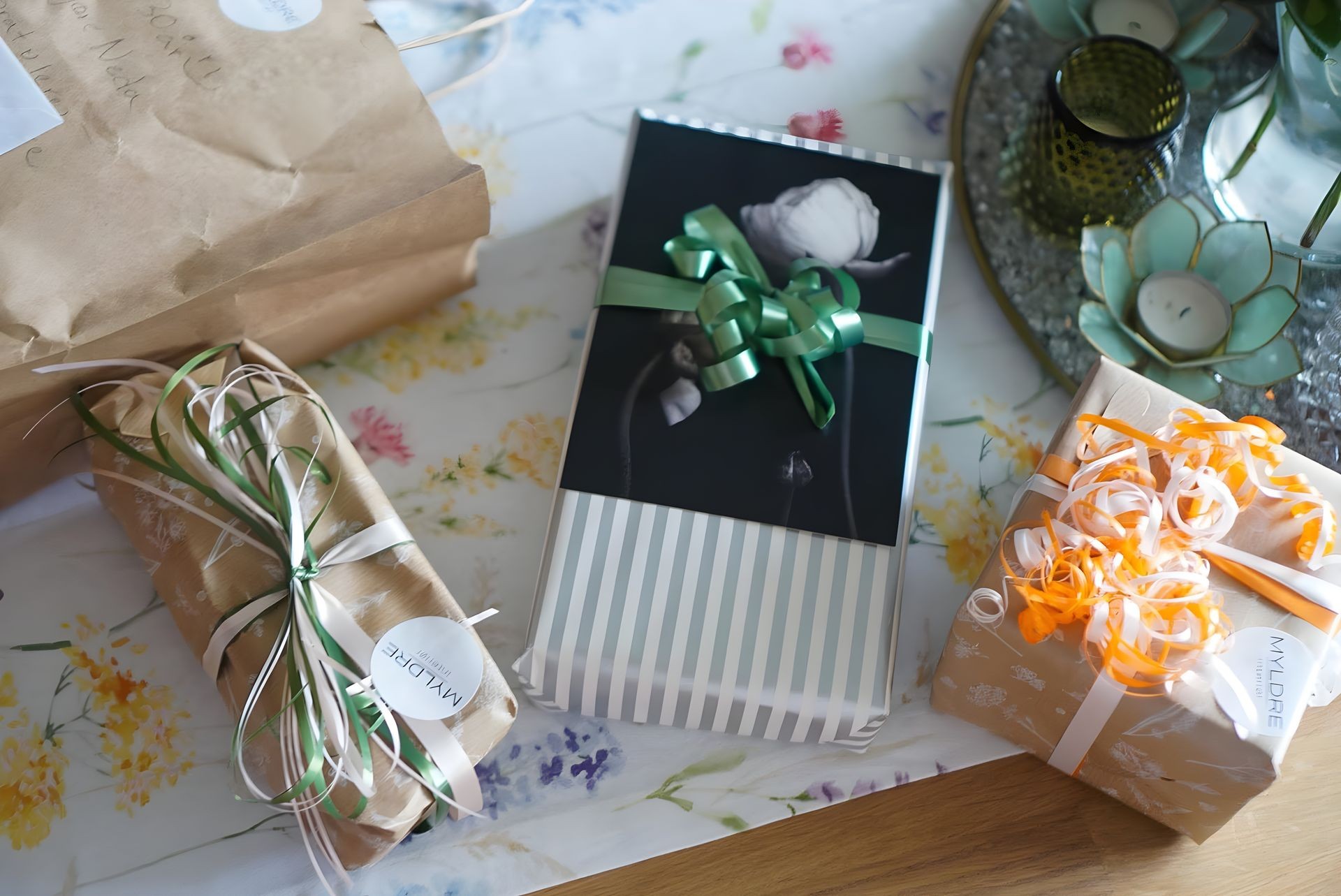 Four wrapped presents with various ribbons on a floral tablecloth, accompanied by candles in decorative holders.