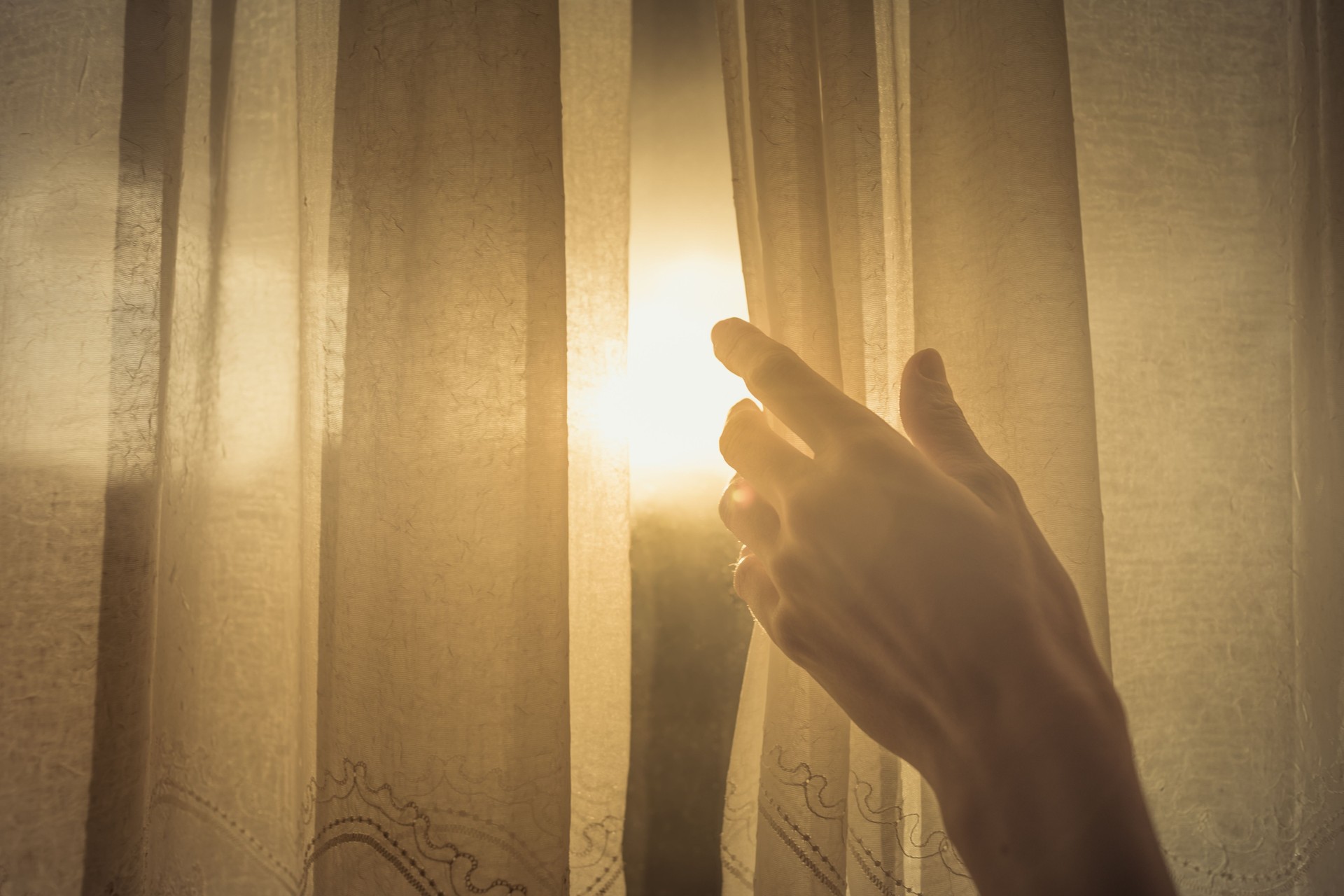Hand opening up window curtain letting the early morning sunshine in.
